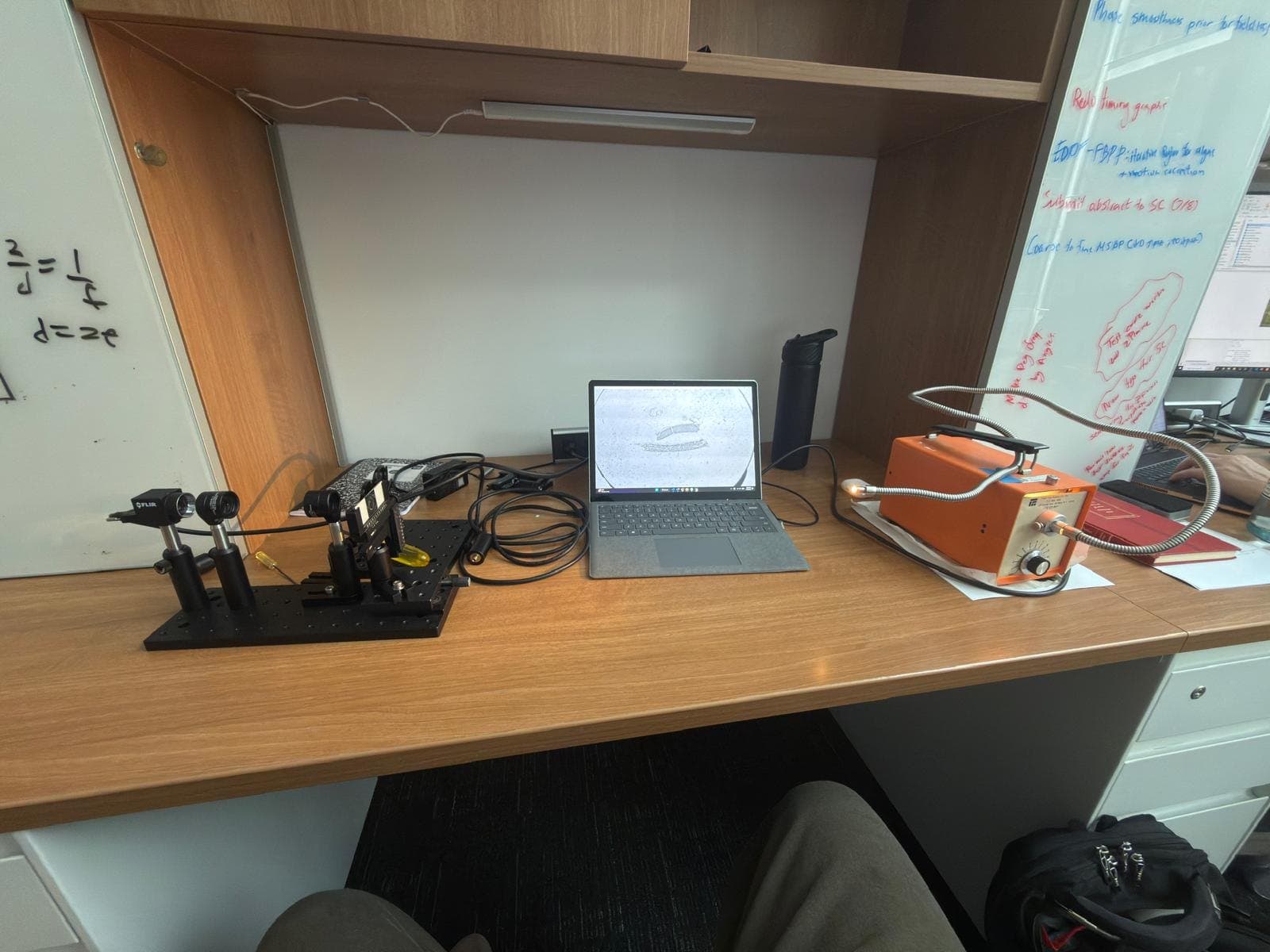 A lab bench with the optical setup, a laptop and an orange instrument in front of a whiteboard covered in equations